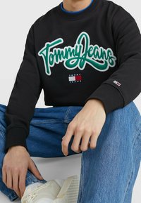 Felpa nera con logo ricamato verde "Tommy Jeans", dettaglio della piccola bandiera, polsini a coste, indossata con jeans blu e sneakers bianche.