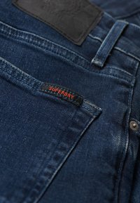 Jean en denim bleu foncé avec un patch en cuir noir à la taille et une petite étiquette "SUPERDRY" en rouge sur la poche avant.