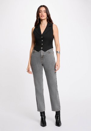 Donna con capelli lunghi e scuri, che indossa un gilet nero senza maniche con bottoni, jeans grigi a vita alta, stivali neri e un braccialetto a polsino d'argento, in piedi su sfondo bianco.