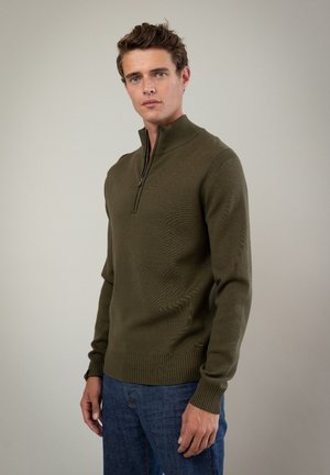 Olivegrøn strikket sweater med en kvart lynlås, ribbet kant og ærmer, med et struktureret mønster, båret over blå denimjeans.