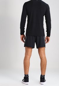 Schwarzes langärmeliges Sportshirt in schlanker Passform, kombiniert mit schwarzen Shorts. Auffällige reflektierende Akzente an den Shorts, glatte Stoffoberfläche.
