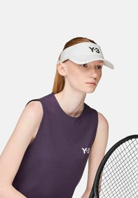 Frau, die eine weiße Y-3-Visiermütze und ein ärmelloses, lila Y-3-Oberteil trägt, mit einem Tennisschläger in der Hand, die konzentriert vor einem schlichten Hintergrund schaut.