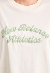 Kremt-skjorte med en hevet, mintgrønn "New Balance Athletics" logo i en rund, kursiv skrift. Bomullsstoff med en myk tekstur.