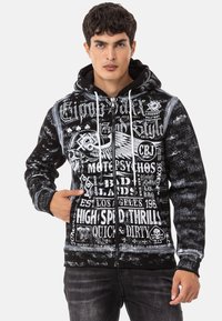 Schwarze Zip-Hoodie mit weißem Grafiktext und Designs, einer Kordelzugkapuze, Fronttaschen und einem strukturierten Stoff mit einem verblassten Muster.