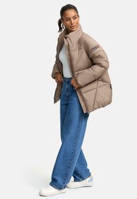  Lichtbruine pufferjas met een hoge kraag, diagonale stikdetails en een zijzak, gecombineerd met hoog getailleerde blauwe jeans en witte sneakers.
