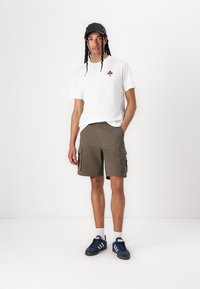 T-shirt blanc à manches courtes avec un petit graphique, shorts cargo olive avec poches latérales, casquette noire et baskets bleues avec accents blancs.