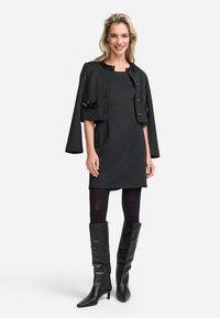 Vestido negro hasta la rodilla con una chaqueta-capa estructurada, cuello adornado y mangas anchas. Acompañado de botas de cuero negras hasta la rodilla.