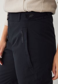 Marineblauwe outdoorbroek met een zachte textuur, voorzien van een zijzak en knoopsluiting, gemarkeerd met borduring van het "67°N Åhkká" logo.