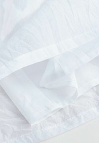 Hvide stoflag med en rynket tekstur, der indeholder glatte og let gennemsigtige sektioner, er pænt foldet med synlige syninger langs kanterne.