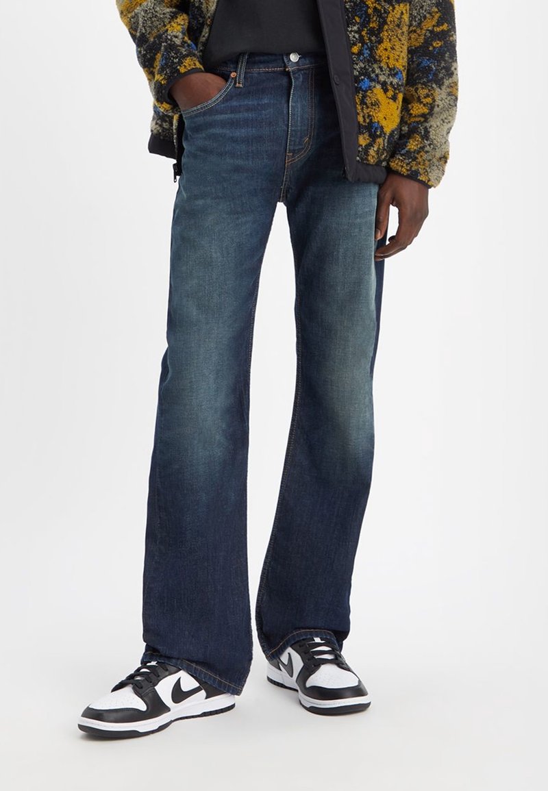 Jean en denim bleu foncé à coupe évasée, avec un léger effet délavé. Associé à des baskets noires et blanches. Aucun logo visible sur le jean.