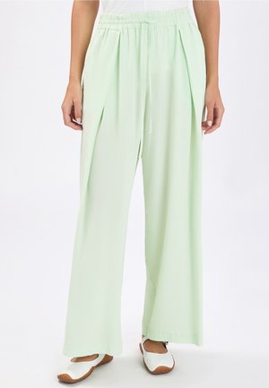 Donna che indossa pantaloni verdi chiaro a gamba larga con una vita elasticizzata e coulisse, abbinati a scarpe bianche basse, in piedi su sfondo bianco.