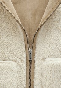 Beige jakke med en blød, fluffy hvid shearling tekstur. Har en central sølvlynlås og to frontlomme.