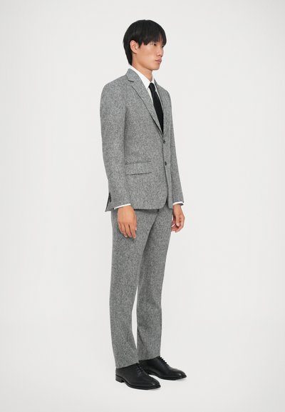 Grauer Tweed-Anzug mit schmaler Passform, ausgestattet mit zwei Knöpfen, Reverskragen und Seitentaschen. Kombiniert mit einem weißen Hemd und schwarzer Krawatte, schwarzen Schuhen.