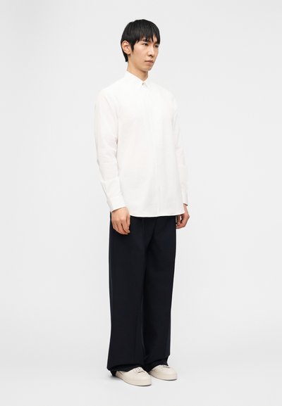 Άνδρας με λευκό πουκάμισο με μακριά μανίκια και κουμπιά, χαλαρά μαύρα παντελόνια και εκρού αθλητικά παπούτσια, στέκεται μπροστά σε απλό φωτεινό φόντο.