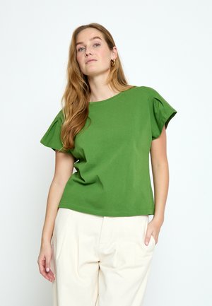 Ženska z dolgimi lasmi, ki nosi zlate obročaste uhane, zeleno kratko majico s plise rameni in krem hlače z visokim pasom, pozira na belem ozadju.