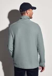 Světlé zelené fleecové pullovrové tričko s vysokým límcem, strukturovaným povrchem a dlouhými rukávy. Nosí se s černou kšiltovkou, zobrazeno zezadu.