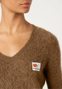 Prune V-kaelusega kampsun, mis on valmistatud karvase materjali jaoks, koos värvilise sildiga, millel on kaks puuvilja illustreerimist ja tekst "LA MAISON AMY alates 2005. aastast."