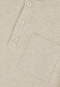 Beige ribbelstof met een henley-kraag, drie crèmekleurige knopen en een voorzak. Zachte textuur, effen patroon zonder zichtbare logo's.