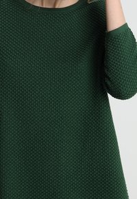 Maglione verde testurizzato con scollo rotondo e maniche a tre quarti con un bordo liscio. Il materiale ha un aspetto lavorato a maglia con un motivo a nido d'ape.