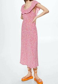 Femme portant une robe midi sans manches rose à fleurs avec un col à volants et des sandales à plateforme orange à brides cheville, sur fond blanc.