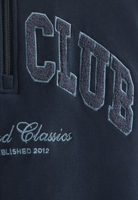 Felpa navy con ricamo in rilievo, morbido grigio, con il testo "CLUB" e "Founded Classics", e dettagli con colletto con zip.