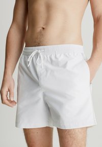 Costume da bagno bianco realizzato in tessuto leggero, dotato di vita elastica, chiusura con cordoncino e tasche laterali. Texture liscia.