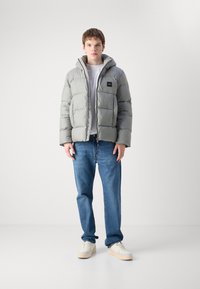 Grå pufferjakke med hætte og lynlås, med en frontlogo-patch, parret med blå jeans og hvide sko.