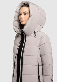 Puffa bunda ve světle šedé barvě, vybavená velkou kapucí, zapínáním na zip vpředu a bočními kapsami. Prošívaná textura s černými zipovými detaily.