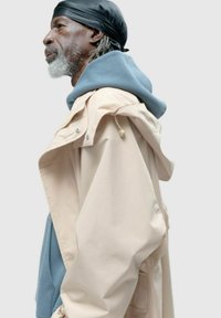 Beżowa wodoodporna kurtka o luźnym kroju, nałożona na jasnoniebieską bluzę z kapturem. Posiada sznurki i zatrzaski.