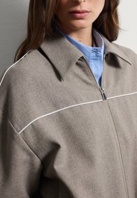 Donna che indossa una giacca marrone con zip e piping bianco sopra una camicia blu chiaro con colletto, mostrata dalla gola fino a metà torace.