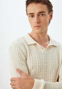 Gestructureerde crèmekleurige sweater met een v-hals en polokraag, met kabelbreipatronen en geribde manchetten. Model poseert met de armen gekruist.