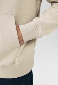 Højre hånd stikkende ind i frontlommen på en beige hættetrøje iført med mørke bukser, der viser ribbede manchetter og kantdetaljer.