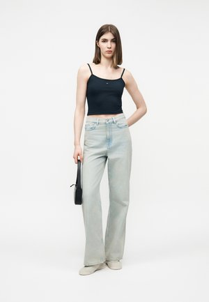 Noor naine seisab, kandes musta lühikest toppi, heleid laiade säärtega pükse, beežid kingad ning käes väikest musta käekotti.