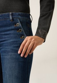 Jeans in denim con un lavaggio scuro, dotati di cinque bottoni metallici sul lato e una manica grigia testurizzata con dettagli in strass.