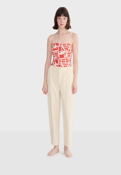 Jeune femme debout, portant un haut sans manches à motifs rouges et blancs, un pantalon crème taille haute et des chaussures plates tissées transparentes.