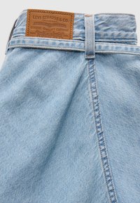 Φωτεινό γαλάζιο denim ύφασμα με υφή στην επιφάνεια, που διαθέτει καφέ δερμάτινο ταμπελάκι με λογότυπο στη μέση μέσης και ορατές ραφές.