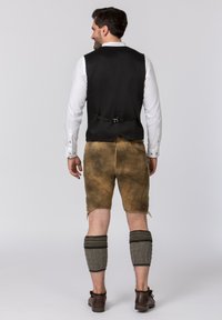 Schwarze Weste mit einem Riemen auf der Rückseite, weißes Hemd, braune Shorts und graue Beinstulpen mit Mustern, kombiniert mit braunen Schuhen.