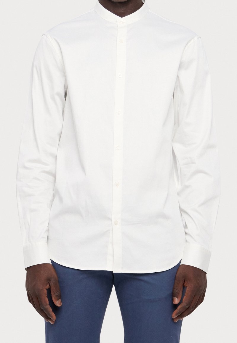 Homme portant une chemise blanche cintrée à boutons avec un col montant et des manches longues, associée à un pantalon bleu.