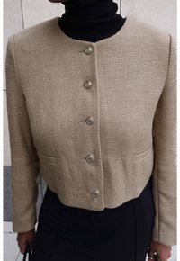 Beige kort jacka med sex metallknappar, strukturerad tyg och två framfickor, bärs över en svart polotröja.