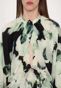 Femme portant un chemisier à col montant avec un motif floral abstrait vert, noir et blanc et une découpe en forme de serrure à l’avant.
