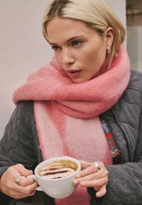 Soft pink wool scarf drapes around the neck, contrasting with a quilted gray jacket. A white ceramic cup holds coffee topped with cocoa design.