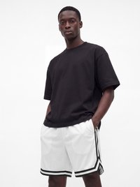 Man wearing a loose black t-shirt and white basketball shorts with black stripes, standing with hands in pockets on a white background.