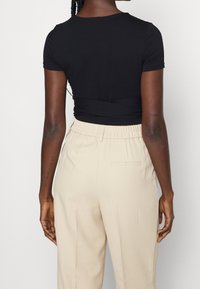 Femme portant un haut noir à manches courtes et un pantalon beige taille haute, debout dos à la caméra sur un fond uni.