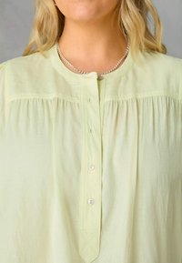 Camicia verde chiaro con bottoni realizzata in tessuto morbido, caratterizzata da un colletto rotondo, dettagli a piccoli plissè e un bottone argentato al colletto.