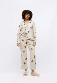 Lys beige pysjamasett med bjørnemønstre, inkludert en skjortetopp med krage og bukser med snøring, som er komplettert med fluffy beige tøfler.
