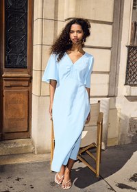 Femme aux cheveux bouclés portant une robe midi bleu clair et des sandales, debout près d'une chaise en bois devant un bâtiment en pierre beige.