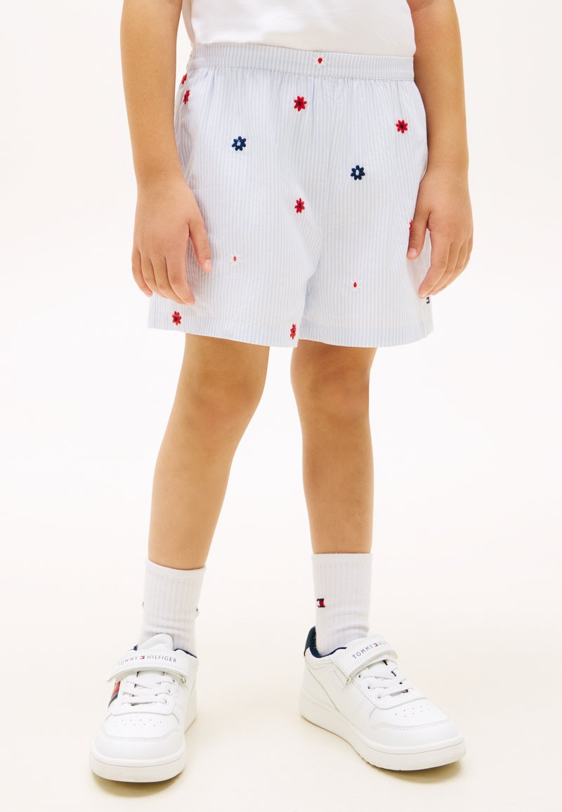 Παιδί που φοράει λευκό σορτς με κεντήματα από κόκκινα και μπλε λουλούδια, λευκές κάλτσες και λευκά αθλητικά παπούτσια με ναυτικό και κόκκινο λογότυπο.