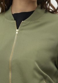 Närbild av en person som bär en olivgrön bomberjacka med en gyllene dragkedja över en svart skjorta.