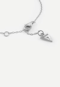 Srebrna bransoletka łańcuchowa z trójkątnym zawieszką oznaczoną napisem "GUESS?" w czarnym kolorze. Posiada zapięcie na karabińczyk oraz okrągłe ogniwa.