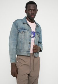 Τζάκετ από τζιν σε ανοιχτό μπλε χρώμα με κλασική γραμμή, διαθέτοντας δύο τσέπες στο στήθος και χάλκινους κουμπιά, φορεμένο πάνω από ένα ροζ μπλουζάκι με γραφικά και μπεζ παντελόνα.